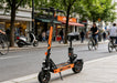 Orange electric scooter on a city street with people cycling and walking.