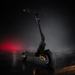 A black electric scooter with gold accents and a large rear tire, photographed in a dark studio setting with atmospheric mist and red lighting on the left.